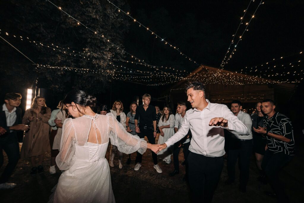 Newly married couple dancing at their wedding with white string lights above them as their friends watch, clap, and enjoy.  