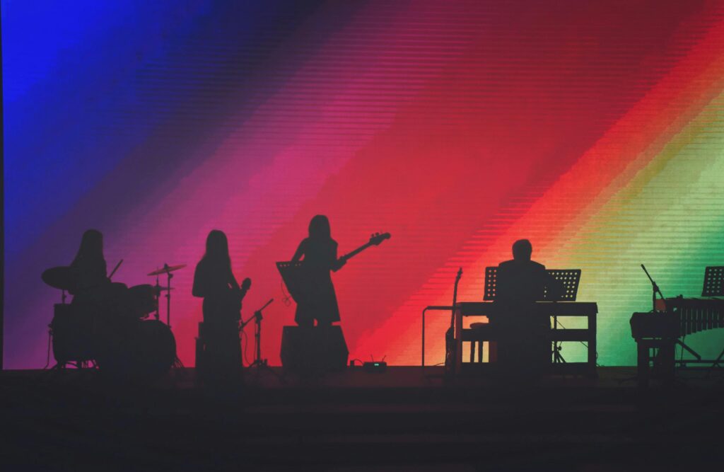 Band performing on stage as silhouettes against a vibrant rainbow-lit backdrop with diagonal beams of blue, red, and green light